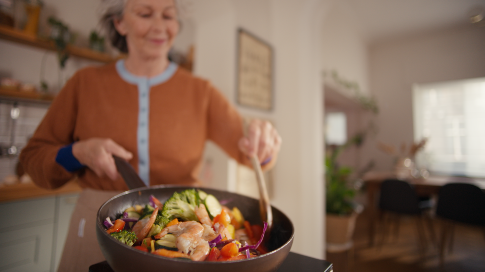 Mosolygó nő, aki egy serpenyőben garnélarákot pirít, és konyhai illatok terjengenek.