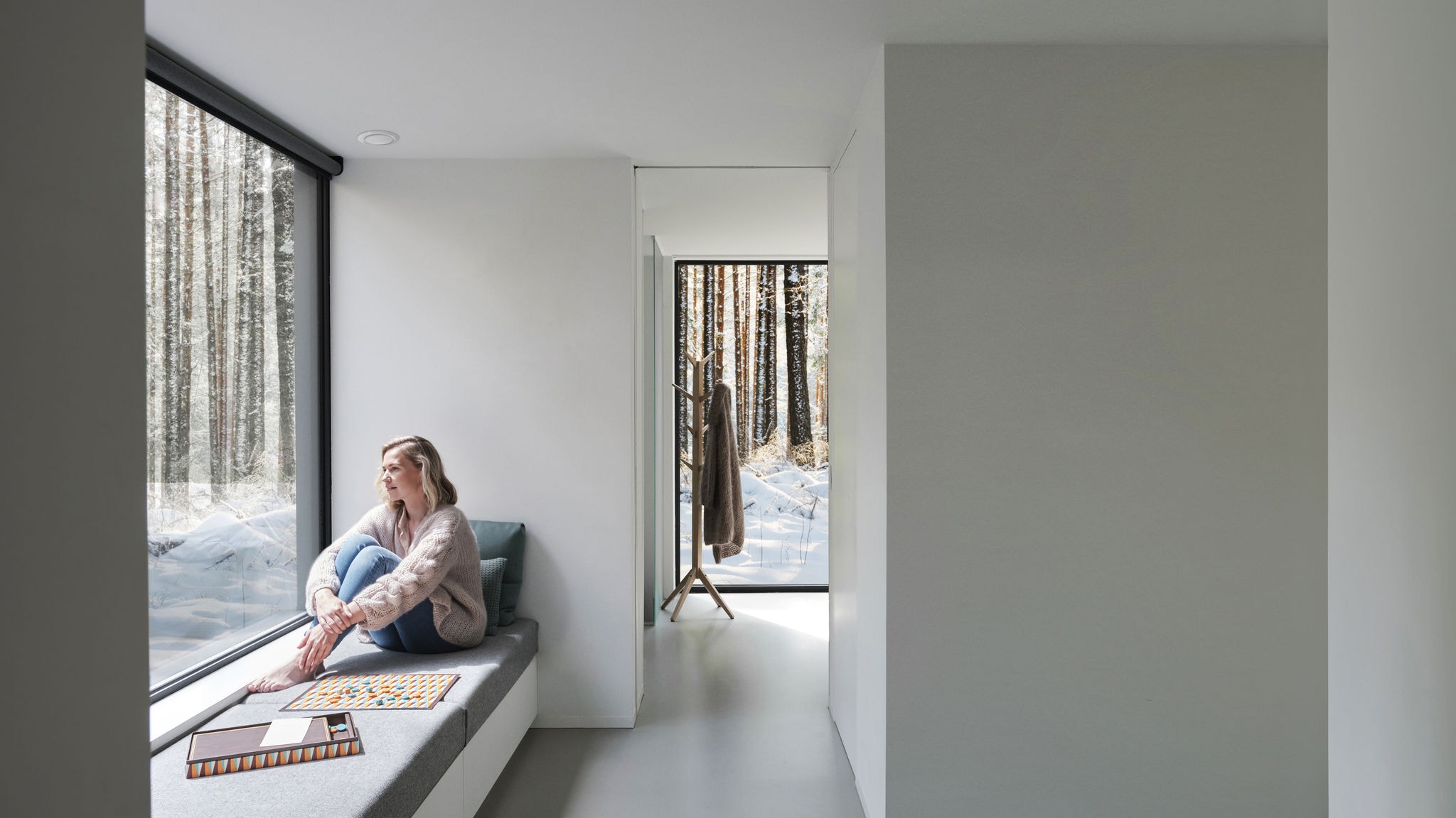 Egy mezítláb ablak előtt ülő nőről készült fotó, aki a fák és a hó felé néz.
