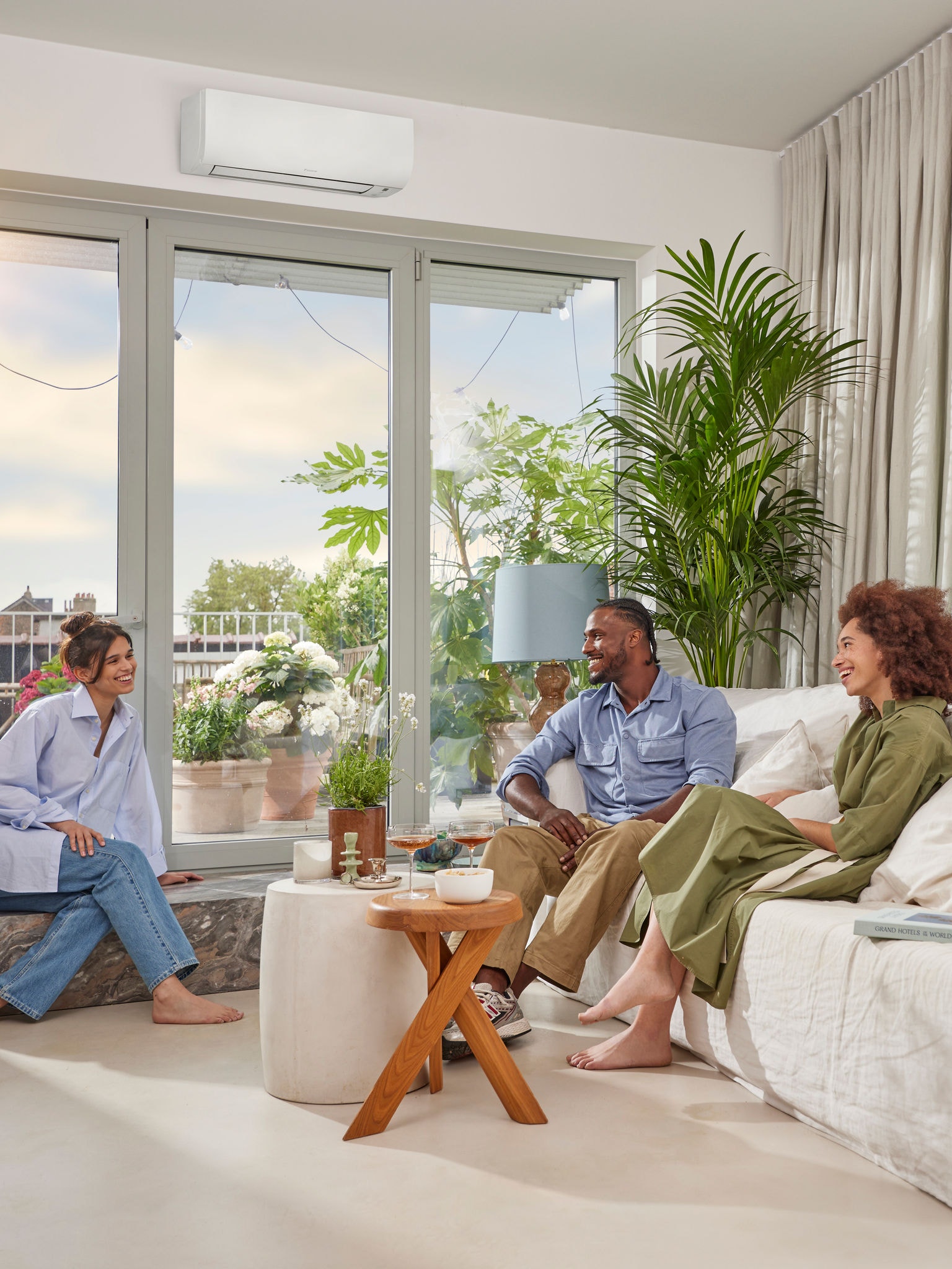 Egy napsütéses napon egy férfi és két nő ülve beszélget egy világos, tágas, fehér falú szobában, ahol egy diszkrét, fehér, oldalfali klíma található egy nagy ablak felett