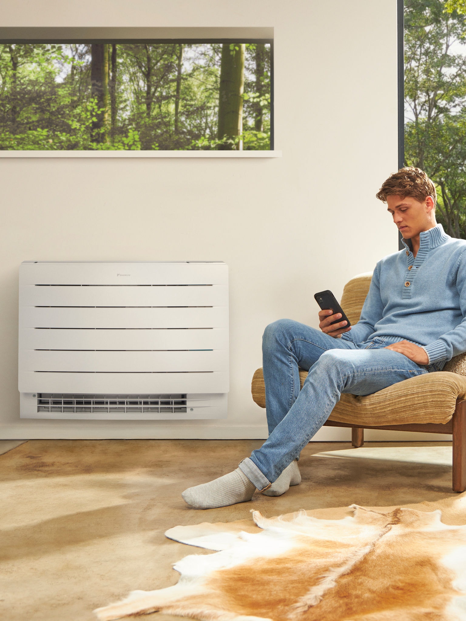 Egy férfi ül egy világos szobában, a telefonját nézi, mögötte pedig a falon egy klíma látható