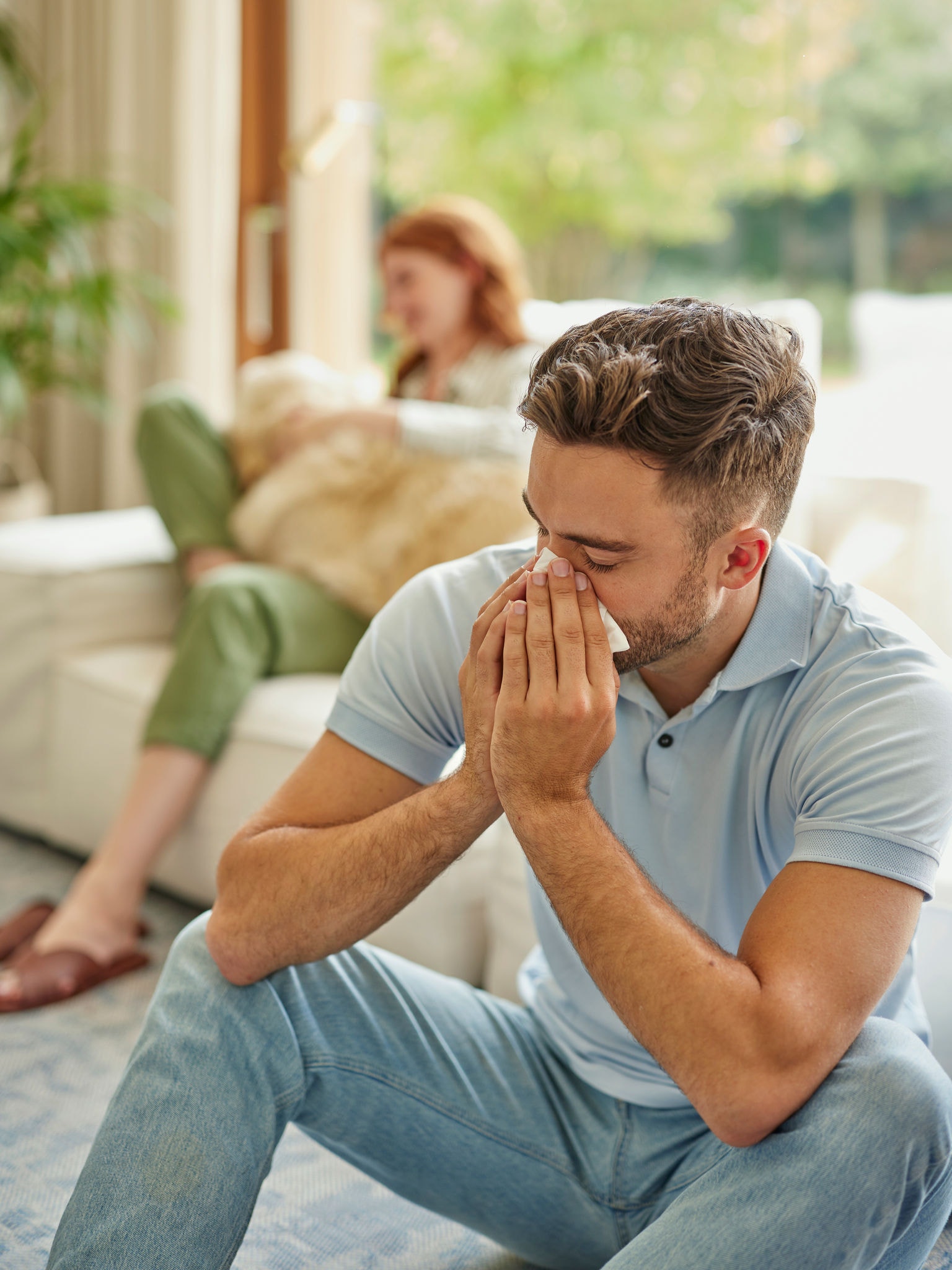 A háttérben puha bútoron ülő nő, ölében egy golden retrieverrel, az előtérben pedig egy férfi tüsszög
