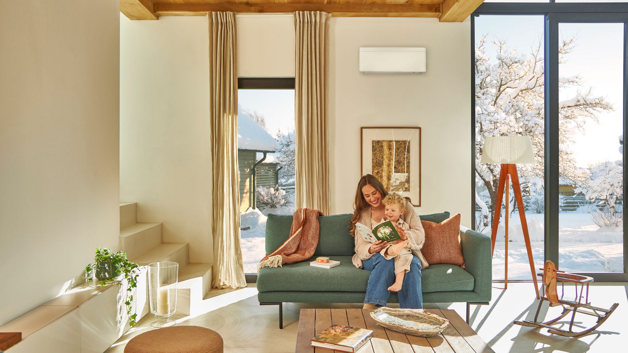 Anya a kanapén ül az oldalfali levegő-levegő hőszivattyú alatt és egy gyermekkönyvet olvas ölében a kisgyermekével, élvezik a benti meleget, míg odakinn havazik